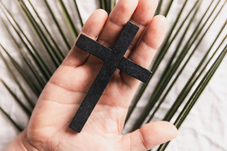 Hand holding wooden cross over white fabric and green palm leaves, symbolizing Christian faith, Palm...