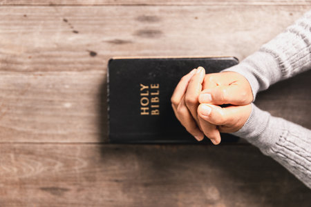 Hands in prayer above Holy Bible on wooden table, symbolizing Christian faith, worship, devotion,...