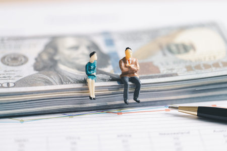 Miniature business people sitting on a stack of US dollar banknotes above a financial chart and pen. Wealth, investment planning, savings and money management concept with copy space.の写真素材