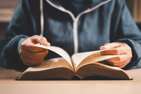 Man reading an open Bible at a desk, turning pages in warm indoor light with shallow depth of field,...