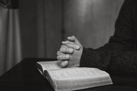 Black and white close-up of praying hands above an open Bible with a wooden cross on a table,...