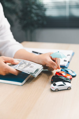 A person holds a stack of banknotes beside a miniature blue umbrella positioned above four toy cars, creating a vertical composition about vehicle-related financial planning.