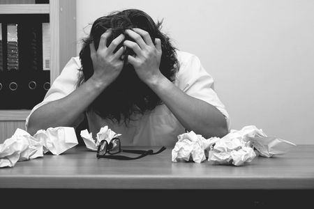 designer holding hands on his long hair while sitting at the desk in very stressful day in the office with many paper trash.の写真素材