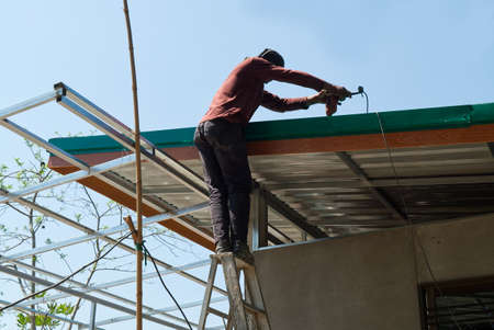 Unsafe Male Roofer Workman Using Electric Screwdriver Install Tile on Roof of New House in the Construction Site with no Protection.の写真素材