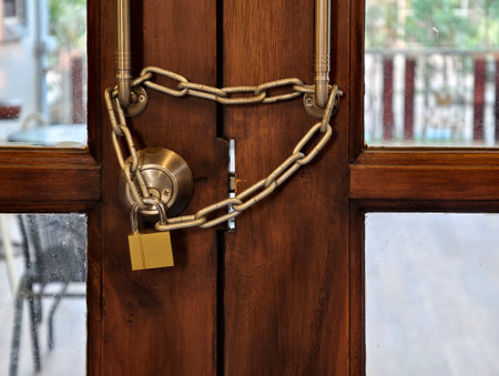 A close-up of a double wooden door tightly secured with a metal chain and brass padlock. This image...