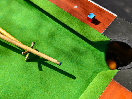Close-up view of a billiard or pool table featuring a wooden cue stick resting on a bridge, a cube of blue chalk, and an orange ball near the corner pocket. The bright green felt surface and natural lighting create a dynamic and nostalgic atmosphere. Ideal for content related to games, sports, leisure, and recreation.の写真素材