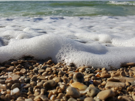 Sea foam surf and shiny multi-colored pebbles. The sea wave has foamed and covers the pebble beach. pebble stones on the sea beach, the rolling waves of the sea with foamの写真素材