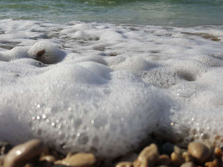 Sea foam surf and shiny multi-colored pebbles. The sea wave has foamed and covers the pebble beach. pebble stones on the sea beach, the rolling waves of the sea with foamの写真素材