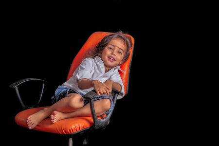a long hair boy sitting on orange chair in black scene in studio to shoot the children's fashion.の写真素材