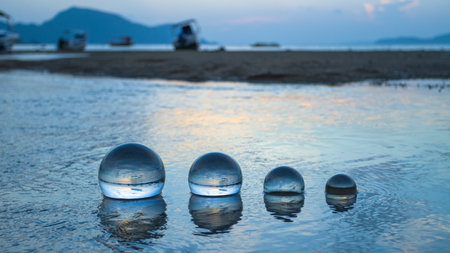 The sun rises within the glass beads arranged in a shallow canal, 
the water gently flows through the orb into the sea. 
A crystal sphere revealing the upside-down sunrise sea view.の写真素材