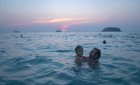 A father carries his child to play in sea water by the beach at sunset. The father is taking the opportunity to bond with his child and enjoy a peaceful evening.の写真素材