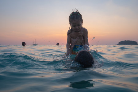 A father carries his child to play in sea water by the beach at sunset. The father is taking the opportunity to bond with his child and enjoy a peaceful evening. The sun is on the palm of his hand.の写真素材