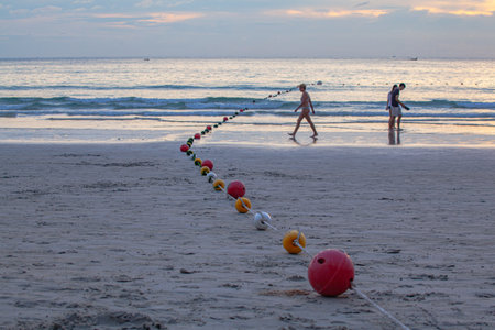 A line of colorful buoys resting on a sandy beach at sunset, gently curving towards the ocean,...