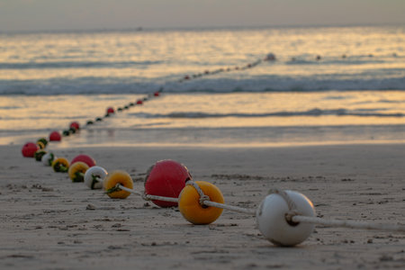 A line of colorful buoys resting on a sandy beach at sunset, gently curving towards the ocean, evoking a sense of calm and the rhythm of coastal life.Buoys create a safety zone for touristsの写真素材