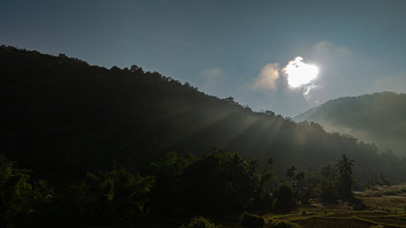 Aerial view of the sun above the mountain peaks shines through the valley. Cloudy skies and surrounding hills enhance the peaceful ambiance of this scenic setting.の写真素材