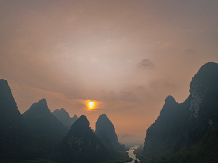 A dramatic sunset view of mountainous landscape with silhouetted karst peaks against an orange sky. A winding river flows through the valley between the mountains.の写真素材