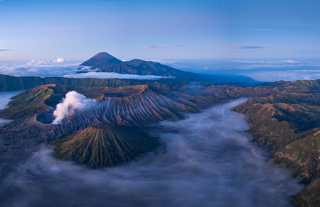 A breathtaking aerial view of Mount Bromo an active volcano in Indonesia, surrounded by mist and dramatic landscapes. The soft morning light highlights the rugged terrain and the rising volcanicの写真素材