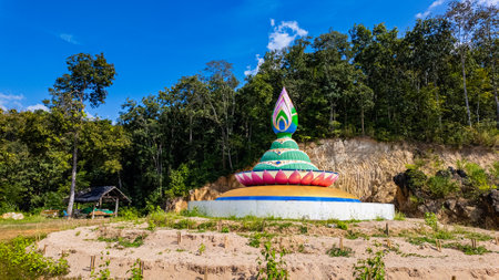 A vibrant stupa in the shape of a lotus, featuring intricate green, pink, and yellow details, set against a bright blue sky with scattered clouds. Surrounded by a serene natural landscape, this spiritual monument stands out as a symbol of faith and tranquility.の写真素材