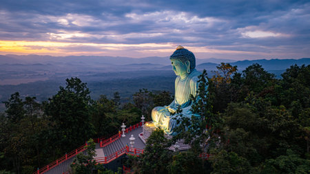 A serene aerial view of a large Buddha statue surrounded by lush forest, with a stunning sunrise casting vibrant hues over a misty mountainous landscape in the background. beautiful sky at sunrise.の写真素材