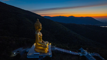 An awe-inspiring view of a big golden Buddha statue atop a hill overlooking a serene valley, captured during the vibrant colors of sunset. aerial view of The biggest golden Buddha at Lopburi Thailandの写真素材