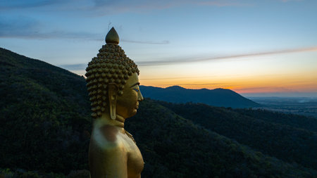 An awe-inspiring view of a big golden Buddha statue atop a hill overlooking a serene valley, captured during the vibrant colors of sunset. aerial view of The biggest golden Buddha at Lopburi Thailandの写真素材
