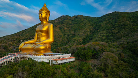 An awe-inspiring view of a big golden Buddha statue atop a hill overlooking a serene valley, captured during the vibrant colors of sunset. aerial view of The biggest golden Buddha at Lopburi Thailandの写真素材