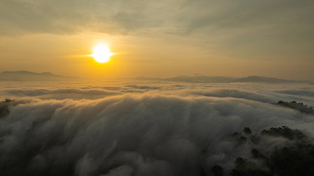 Breathtaking aerial view of the sun rising over a mountain valley covered in a dense sea of rolling clouds. The golden light illuminates the mist, creating a dramatic and ethereal atmosphere at dawnの写真素材