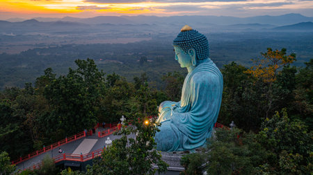 A serene aerial view of a large Buddha statue surrounded by lush forest, with a stunning sunrise casting vibrant hues over a misty mountainous landscape in the background. beautiful sky at sunrise.の写真素材