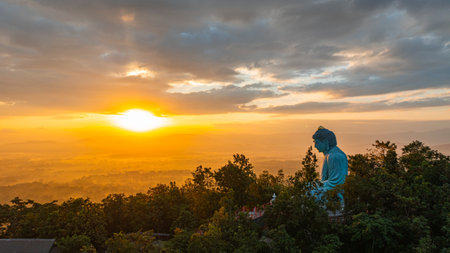 A serene aerial view of a large Buddha statue surrounded by lush forest, with a stunning sunrise casting vibrant hues over a misty mountainous landscape in the background. beautiful sky at sunrise.の写真素材