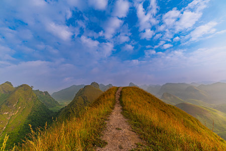 A small path on the ridge, white fluffy clouds in the blue sky float above the rugged mountains. lush vegetation, perfect for nature lovers and outdoor adventure themes. Beautiful sceneryの写真素材