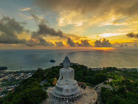 A breathtaking aerial view of the iconic Big Buddha statue overlooking the Andaman Sea in Phuket, Thailand, captured during a vibrant sunset. The golden sky reflects on the water. beautiful sunsetの写真素材