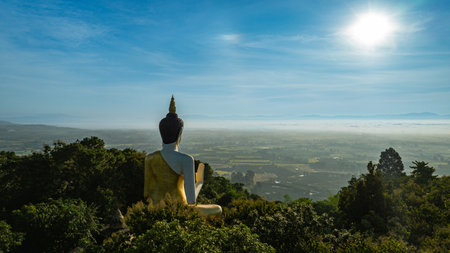 Aerial view of A golden Buddha statue surrounded by lush green trees, set against a stunning backdrop of rolling mountains and a clear blue sky. the harmony of spirituality and nature.の写真素材