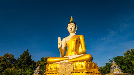 A majestic golden Buddha statue surrounded by lush green trees, set against a stunning backdrop of rolling mountains and a clear blue sky. This serene landscape captures the harmony of spiritualityの写真素材
