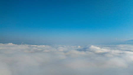 A serene view of the early morning sun rising above a vast sea of clouds, with distant mountain peaks faintly visible on the horizon, creating a peaceful and ethereal landscape. Flying over the cloudの写真素材