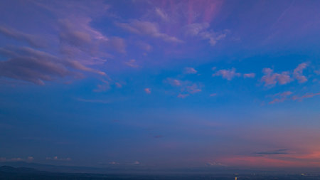 A breathtaking aerial panoramic view of a vibrant sunset illuminating the sky above a lush green hilltop with a small structure, surrounded by scattered clouds and distant landscapes.の写真素材