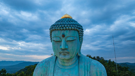 A serene aerial view of a large Buddha statue surrounded by lush forest, with a stunning sunrise...