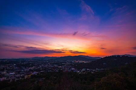 A scenic view of a town nestled among lush green mountains at sunset, with a temple structure perched on the hillside and a vibrant orange sky creating a warm, tranquil atmosphere.の写真素材