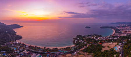 A breathtaking aerial view of Kata Beach in Phuket, Thailand, captured during sunset. The image showcases the coastline, small islands, lush green hills, and the vibrant cityscape under a bright skyの写真素材
