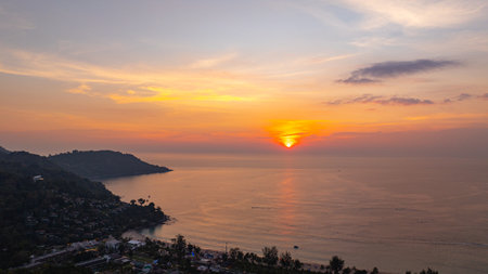 A breathtaking aerial view of Kata Beach in Phuket, Thailand, captured during sunset. The image showcases the coastline, small islands, lush green hills, and the vibrant cityscape under a bright skyの写真素材