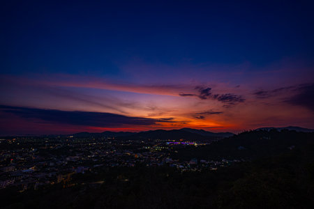 A scenic view of a town nestled among lush green mountains at sunset, with a temple structure perched on the hillside and a vibrant orange sky creating a warm, tranquil atmosphere.の写真素材