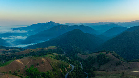 A breathtaking aerial view of a mist-covered forest at sunrise. The dramatic cloudscape and soft fog create a serene and mystical atmosphere. Misty Sunrise Over Forested Hills with layers of mountainの写真素材