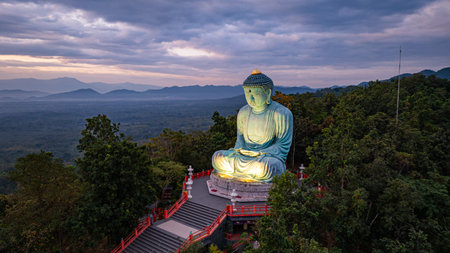 A serene aerial view of a large Buddha statue surrounded by lush forest, with a stunning sunrise casting vibrant hues over a misty mountainous landscape in the background. beautiful sky at sunrise.の写真素材