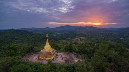 A stunning aerial view of a golden pagoda nestled amidst lush green hills during a vibrant sunset, with dramatic clouds and soft sunlight casting a serene glow over the landscape.の写真素材