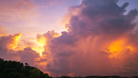 A vivid and dynamic sky captures towering storm clouds illuminated by the warm hues of a setting sun, creating a striking contrast between light and shadow above a darkened forested landscape.の写真素材