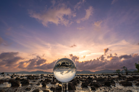 A mesmerizing sunset over a rocky shoreline captured through a glass crystal ball, creating a surreal inverted reflection of the seascape beneath vibrant, colorful skies.の写真素材