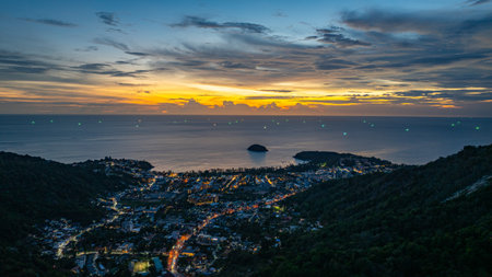 A breathtaking sunset paints the sky in vivid hues of orange and blue over a coastal town, with lush green hills framing the scene. A distant hilltop structure adds a serene tranquil landscape.の写真素材