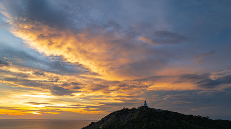 A breathtaking sunset paints the sky in vivid hues of orange and blue over a coastal town, with lush green hills framing the scene. A distant hilltop structure adds a serene tranquil landscape.の写真素材