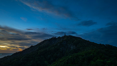 A breathtaking aerial view of the iconic Big Buddha statue overlooking the Andaman Sea in Phuket, Thailand, captured during a vibrant sunset. The golden sky reflects on the water.の写真素材