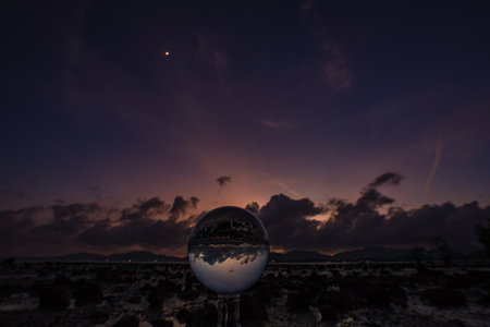 A mesmerizing sunset over a rocky shoreline captured through a glass crystal ball, creating a surreal inverted reflection of the seascape beneath vibrant, colorful skies.の写真素材