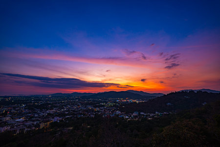 A scenic view of a town nestled among lush green mountains at sunset, with a temple structure perched on the hillside and a vibrant orange sky creating a warm, tranquil atmosphere.の写真素材